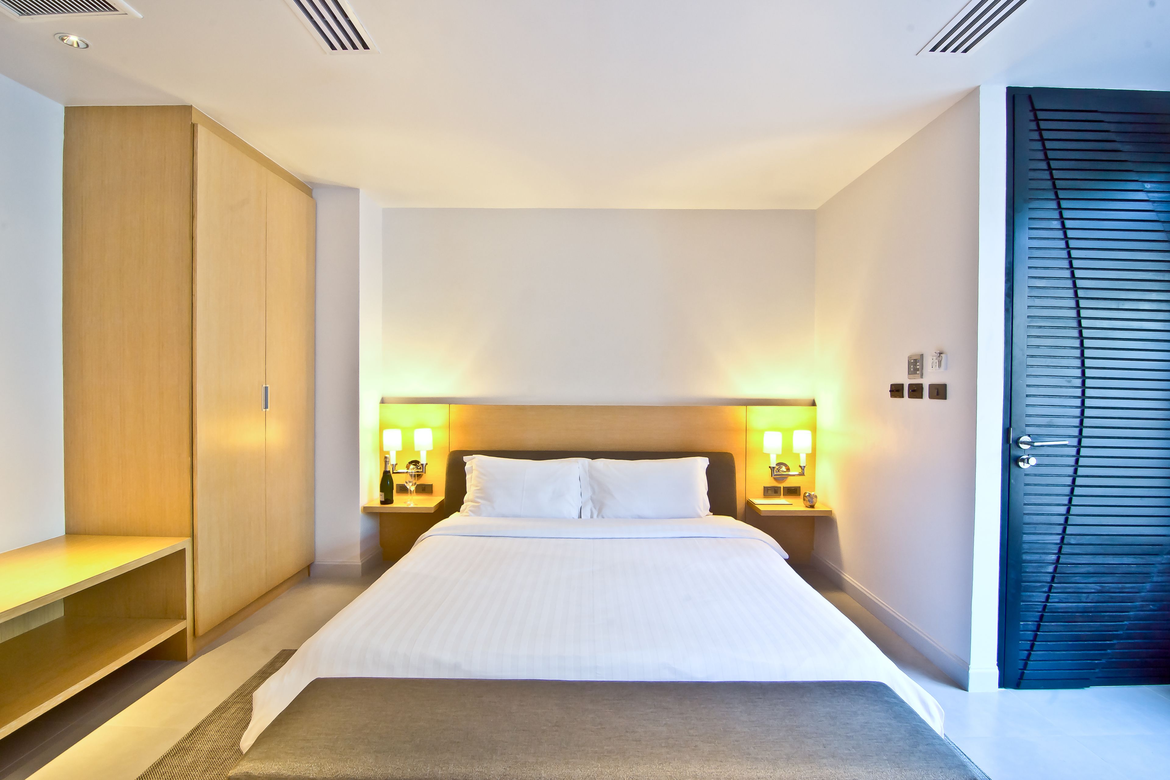 A modern hotel room with a large bed, wooden wardrobe, bedside lamps, and a dark blue door.