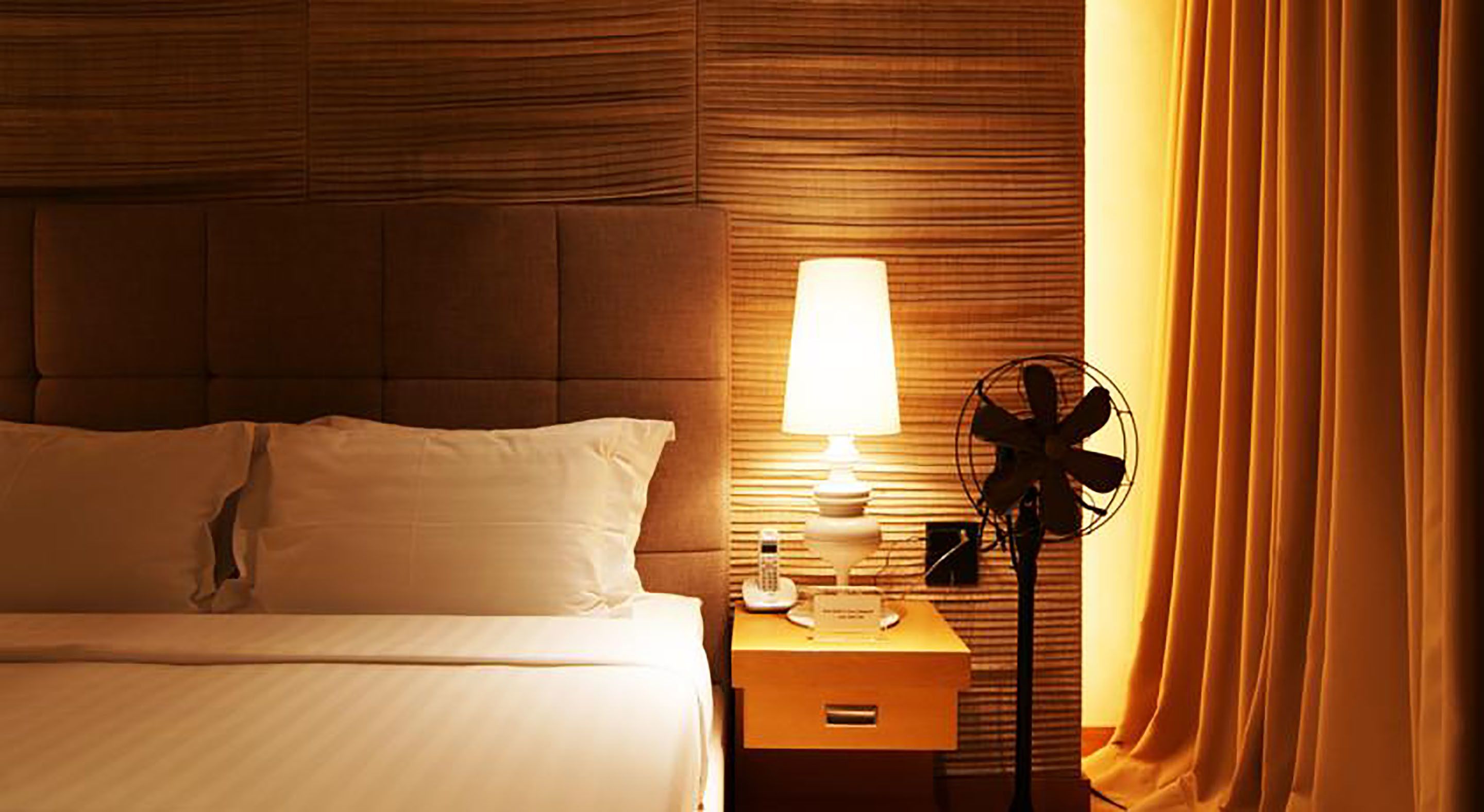 A hotel room with a bed, a bedside table with a lamp, a fan, and a nightstand.