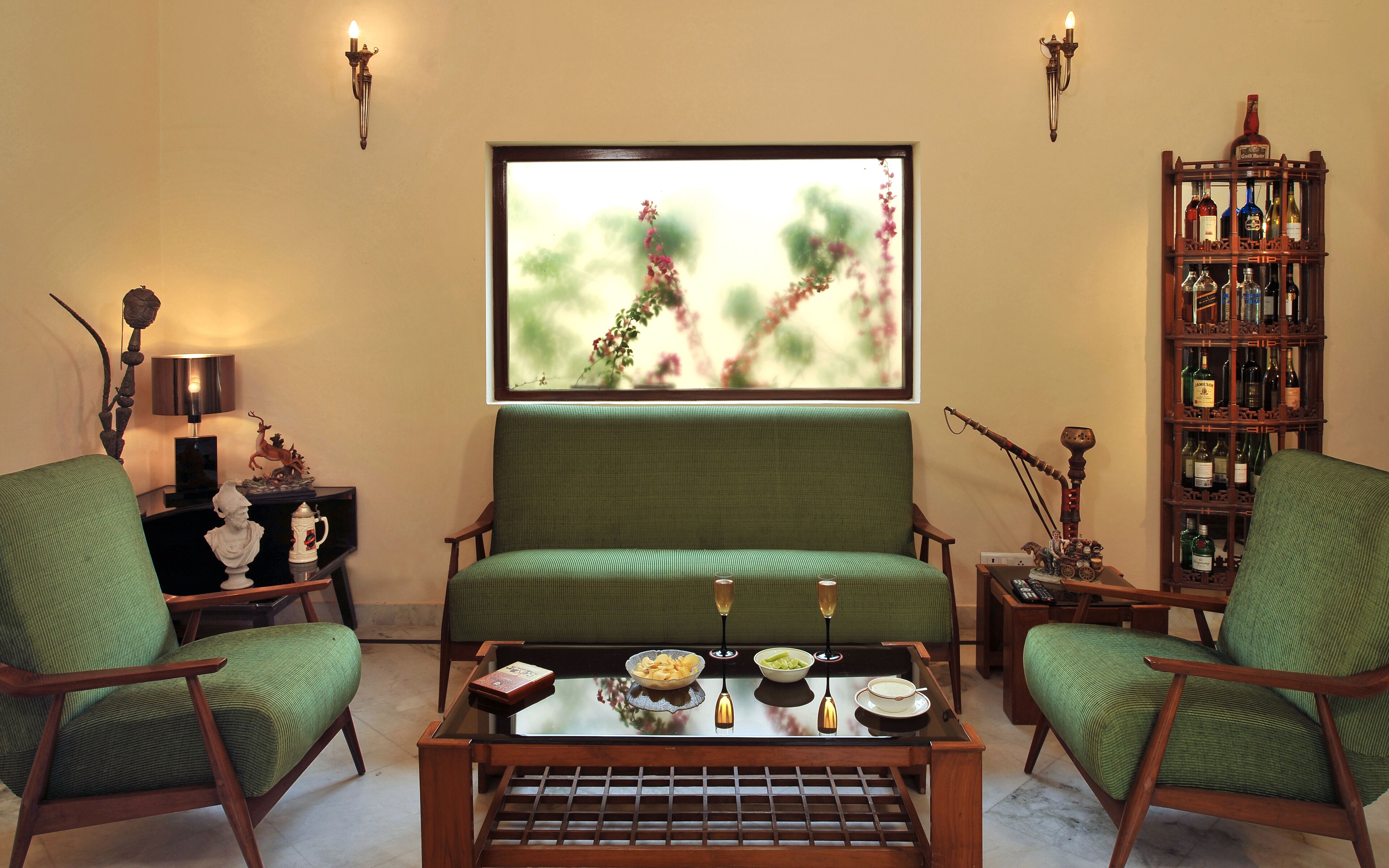A living room with a green sofa, two green armchairs, a coffee table with bowls and glasses, a bookshelf with bottles, and a painting on the wall.
