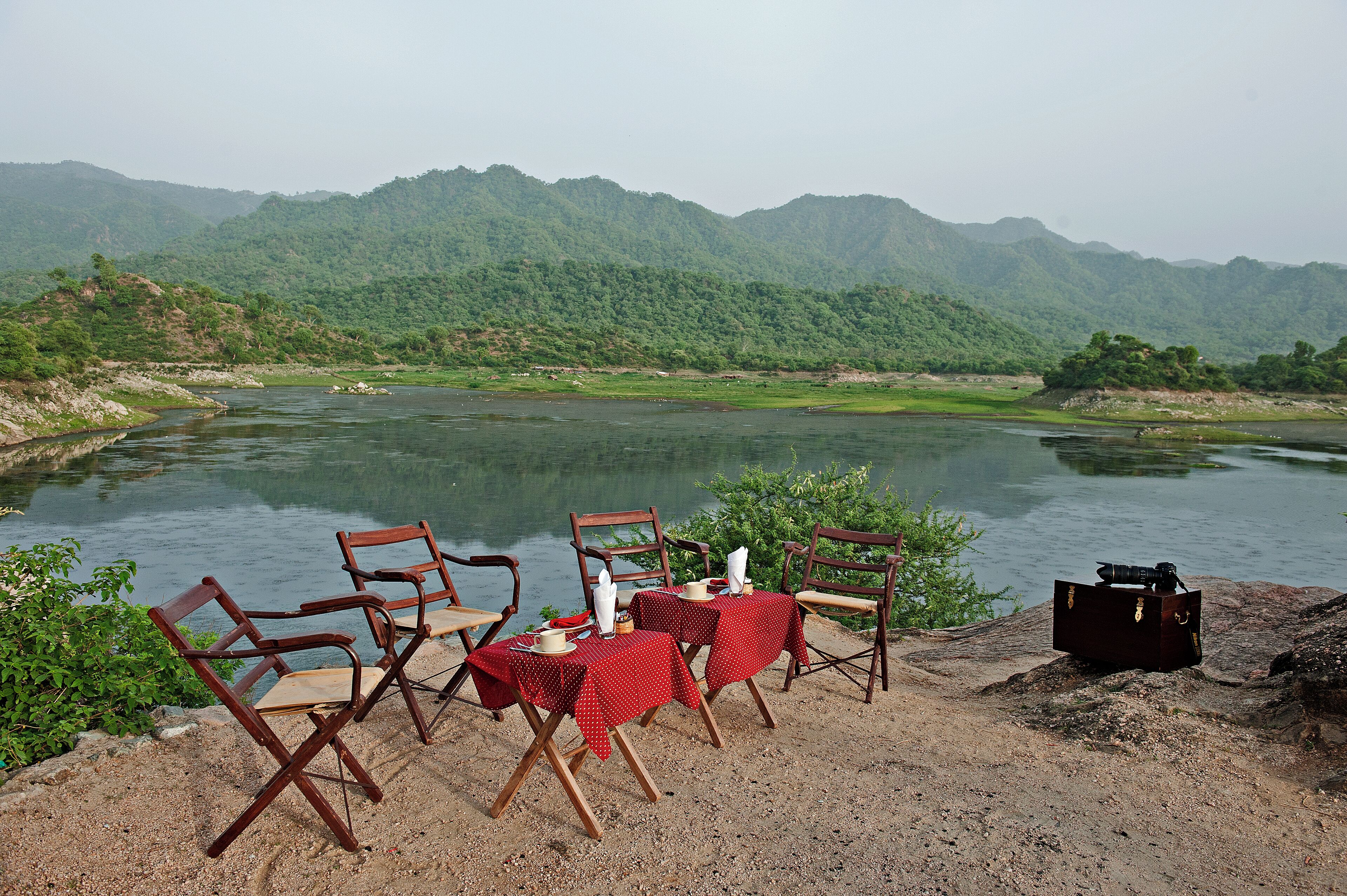 Un espace aménagé pour les repas, avec des chaises et des tables, situé au bord d’une rivière et entouré de montagnes.