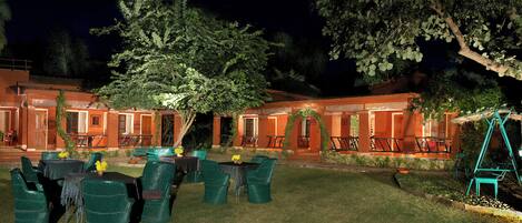 Un patio con mesas al aire libre, un árbol grande y un edificio con varias puertas y ventanas.