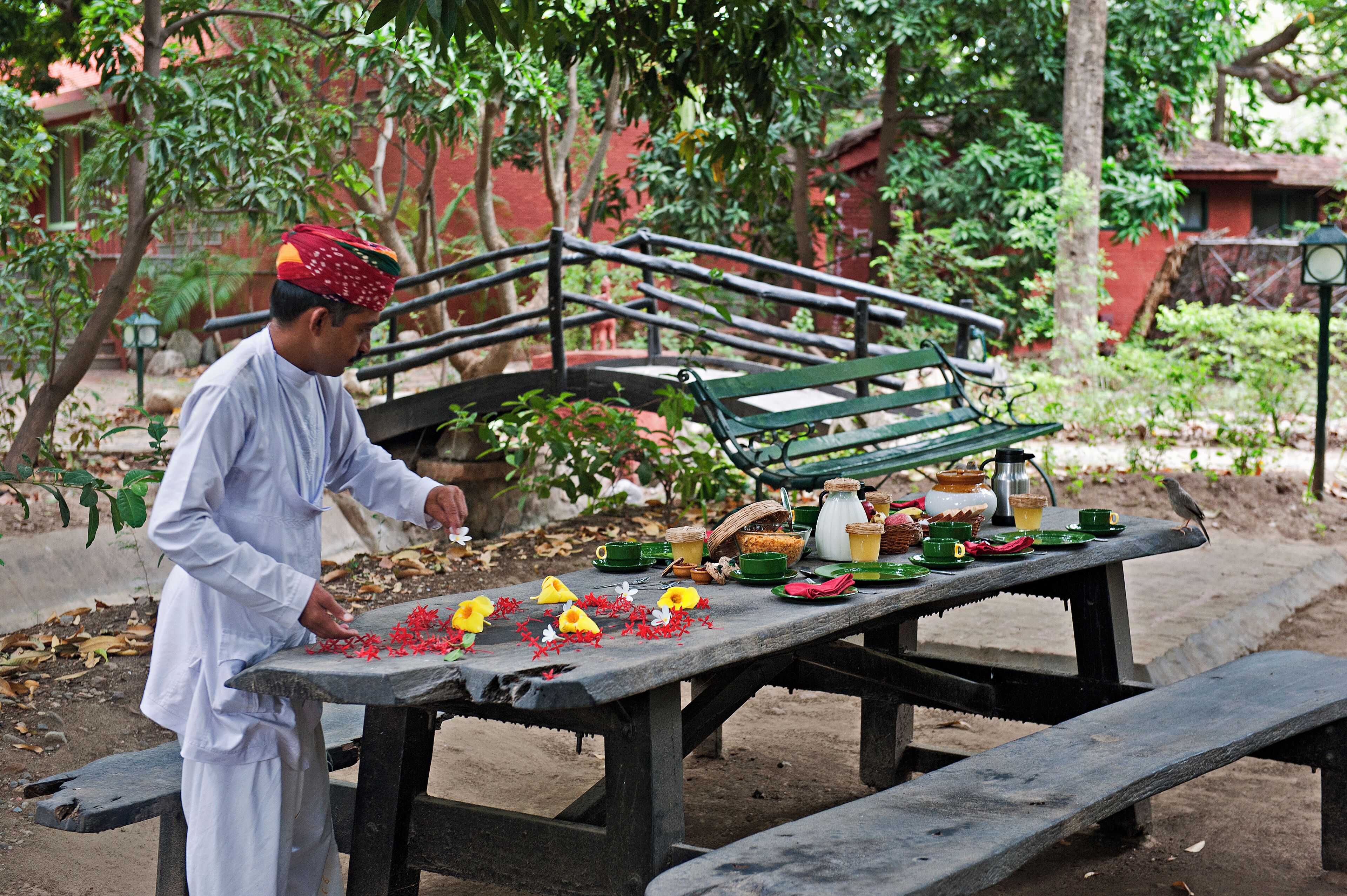 A man arranging flowers on a wooden table outdoors, with a bench and a clock post in the background.
