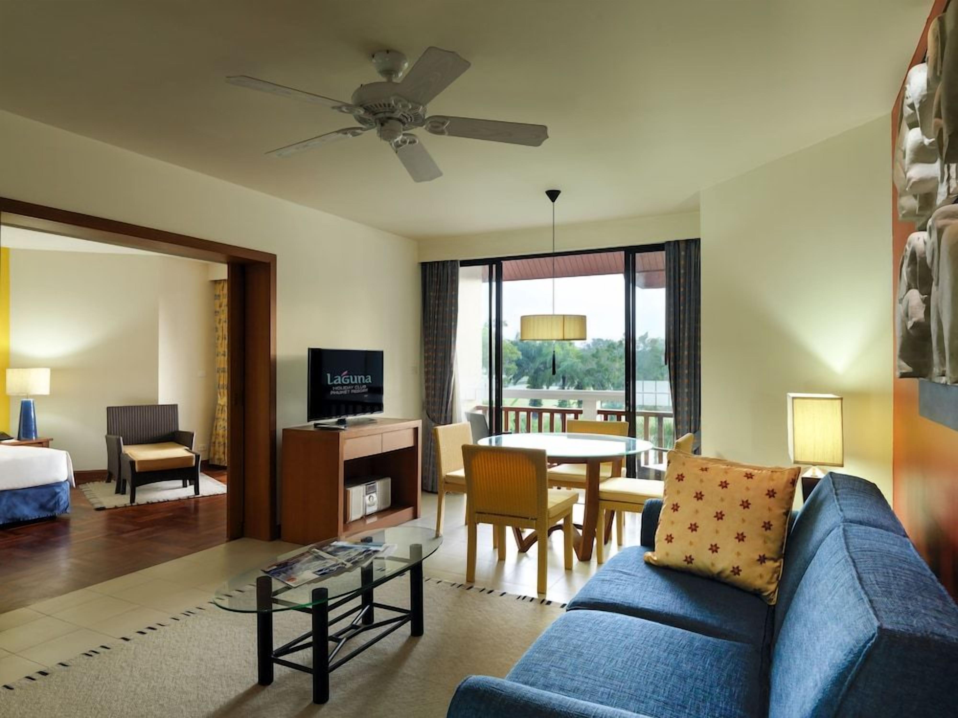 A hotel room with a bed, a TV, a desk, a dining area, and a balcony with a view.