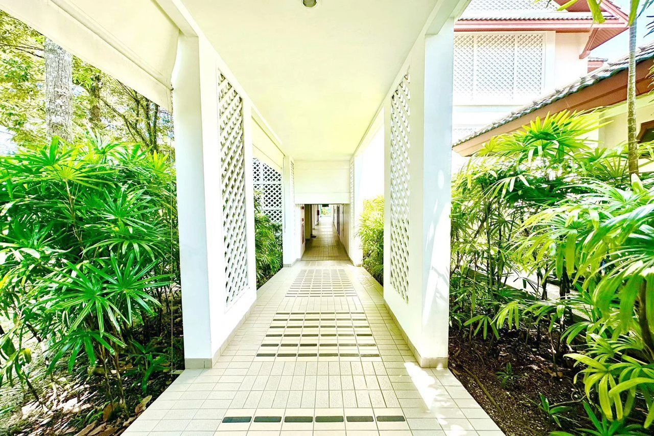 A covered corridor with tiled flooring and lattice panels on the sides, surrounded by lush greenery.