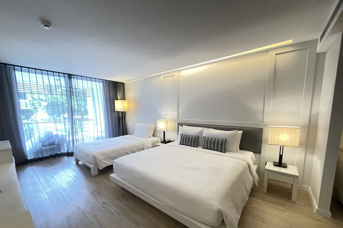 A hotel room with two beds, a large window with curtains, and a bedside table with a lamp.