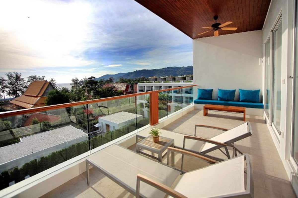 A balcony with a wooden ceiling, a glass railing, a sofa with blue cushions, and a view of the surrounding landscape.