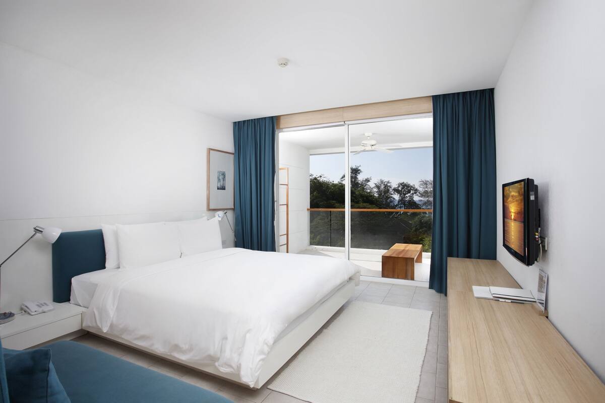 A modern hotel room with a large bed, a desk, a flat-screen TV, and a sliding glass door leading to a balcony.