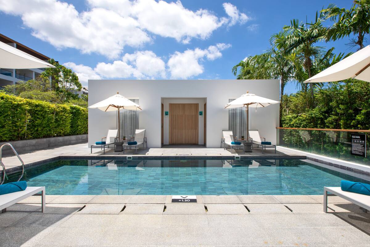 A swimming pool with lounge chairs and umbrellas, surrounded by greenery and a clear sky.