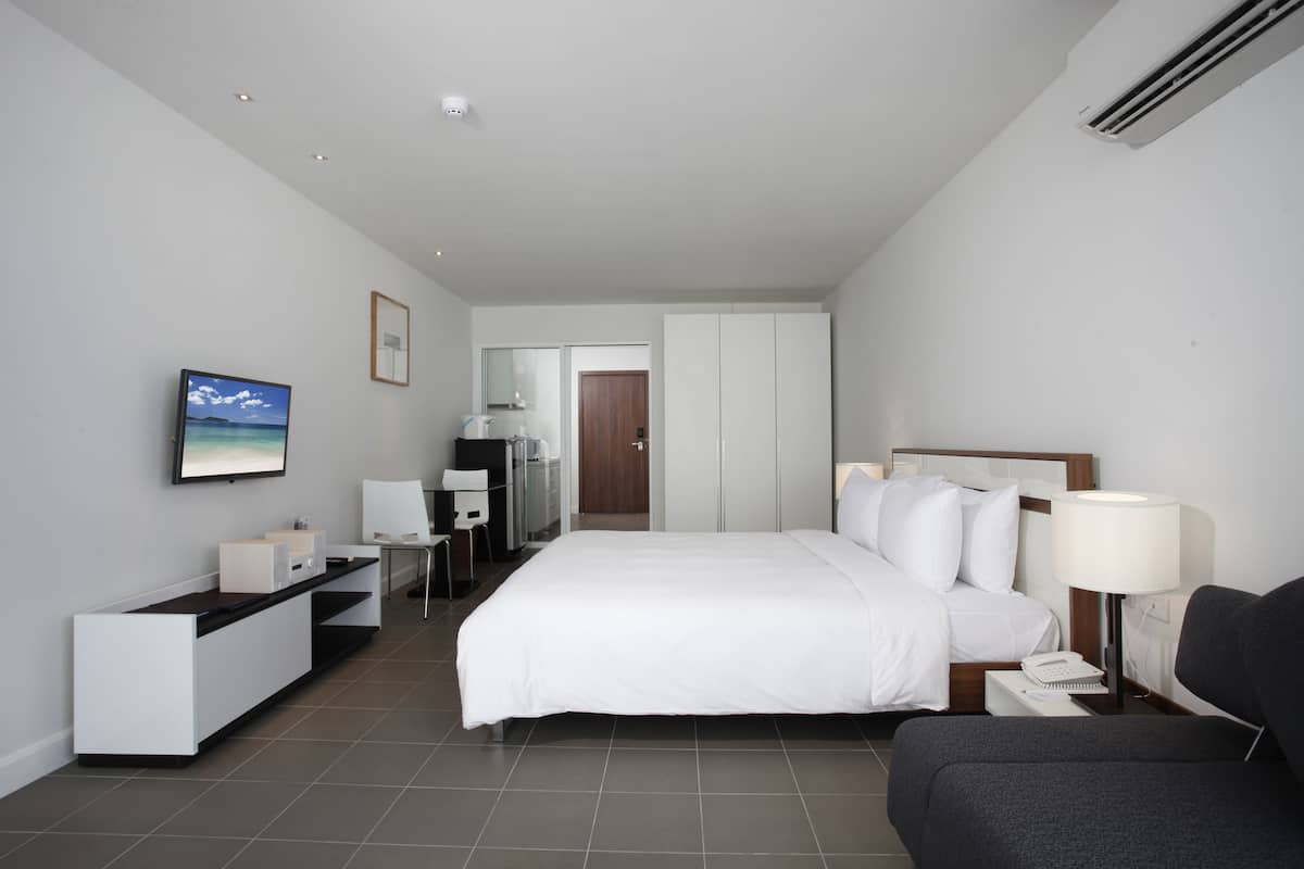 A modern hotel room with a large bed, a desk, a chair, a TV, and a bedside table with a lamp.