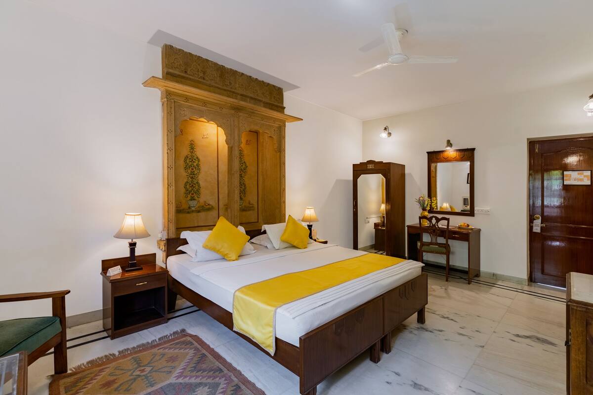 A hotel room with a large bed, two bedside tables with lamps, a wooden wardrobe, a desk with a chair, a mirror, and a door.