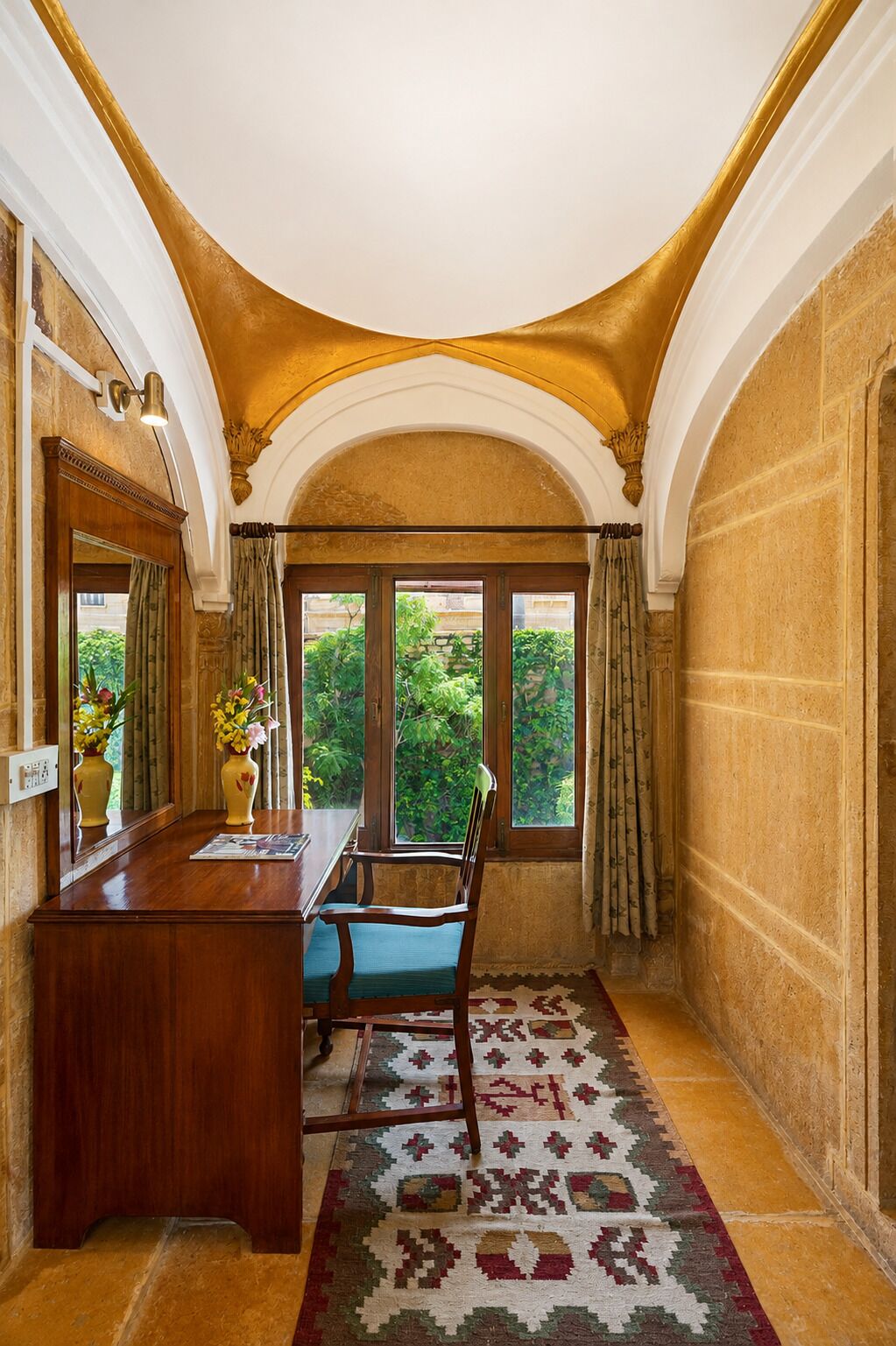 A room with a wooden desk, a chair, a patterned rug, and a view of greenery through large windows.
