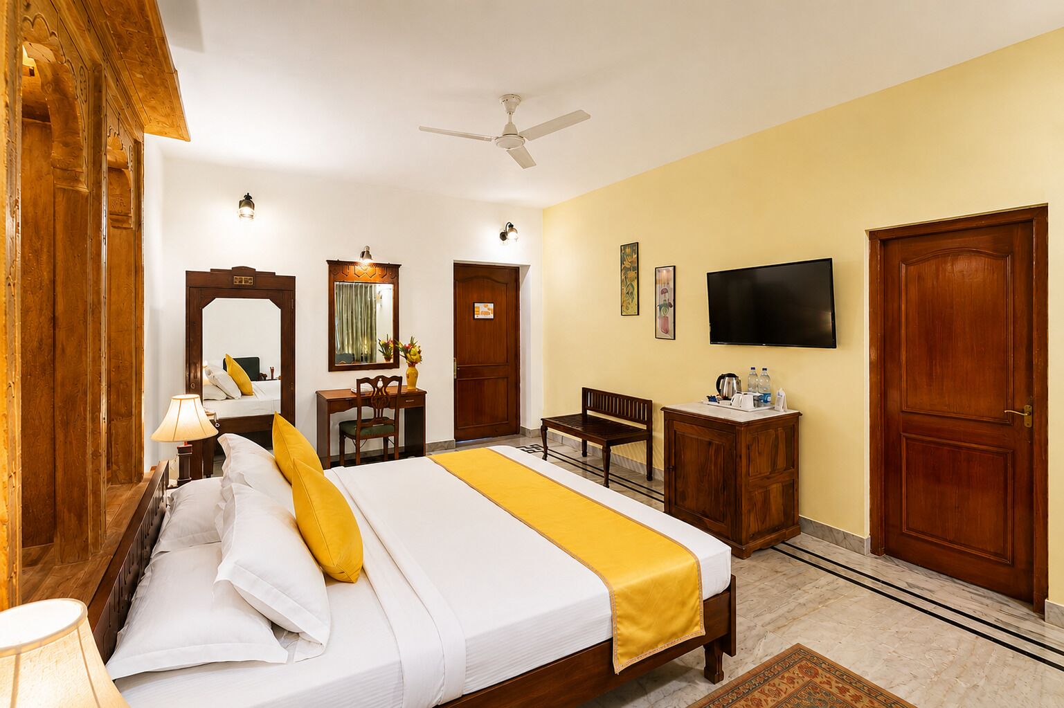 Habitación de hotel con cama, mesita de noche, lámpara, escritorio, silla, televisor, armario y ventilador de techo.