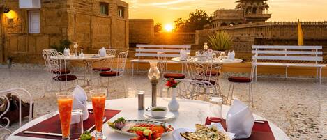 An outdoor dining area with tables and chairs, a sunset view, and a historic building in the background.