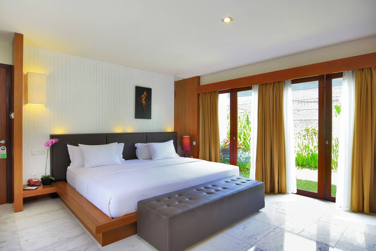 Quarto moderno com uma cama grande, um banquinho e vista para o verde pela janela.