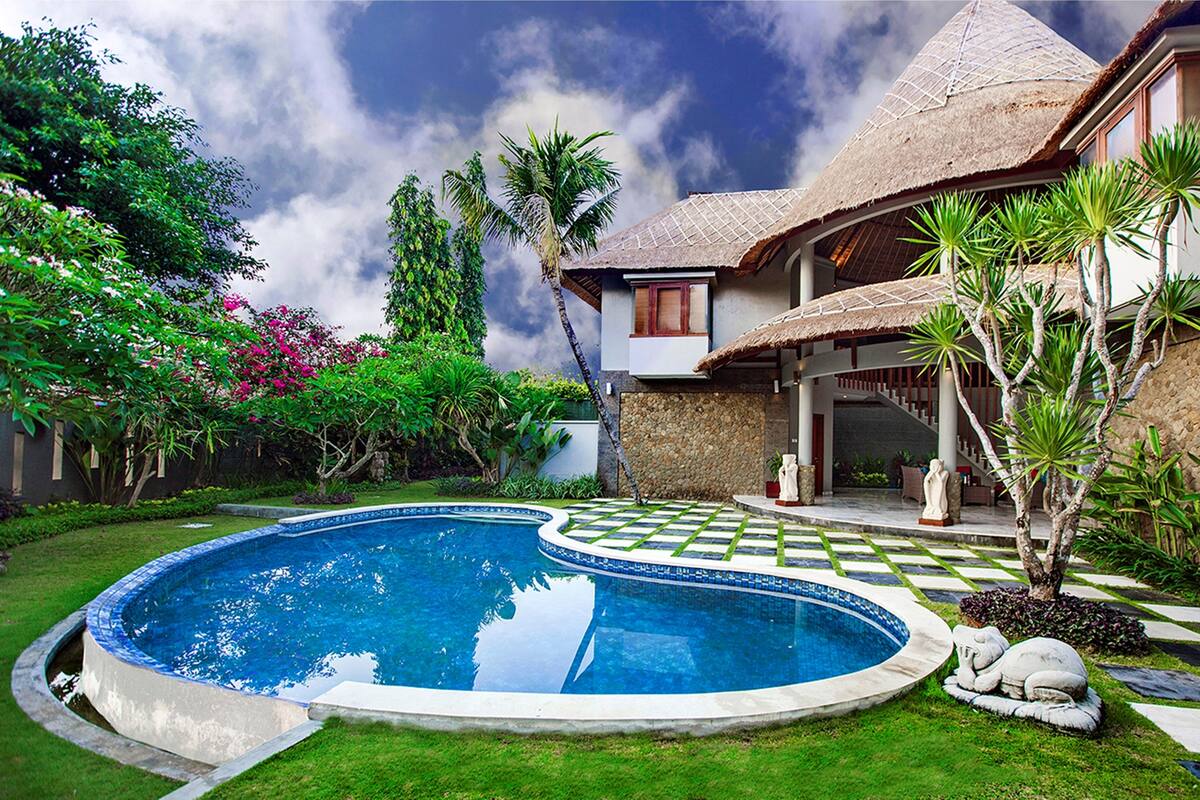 Piscina rodeada por um caminho quadriculado, vegetação exuberante e uma casa com telhado de palha.