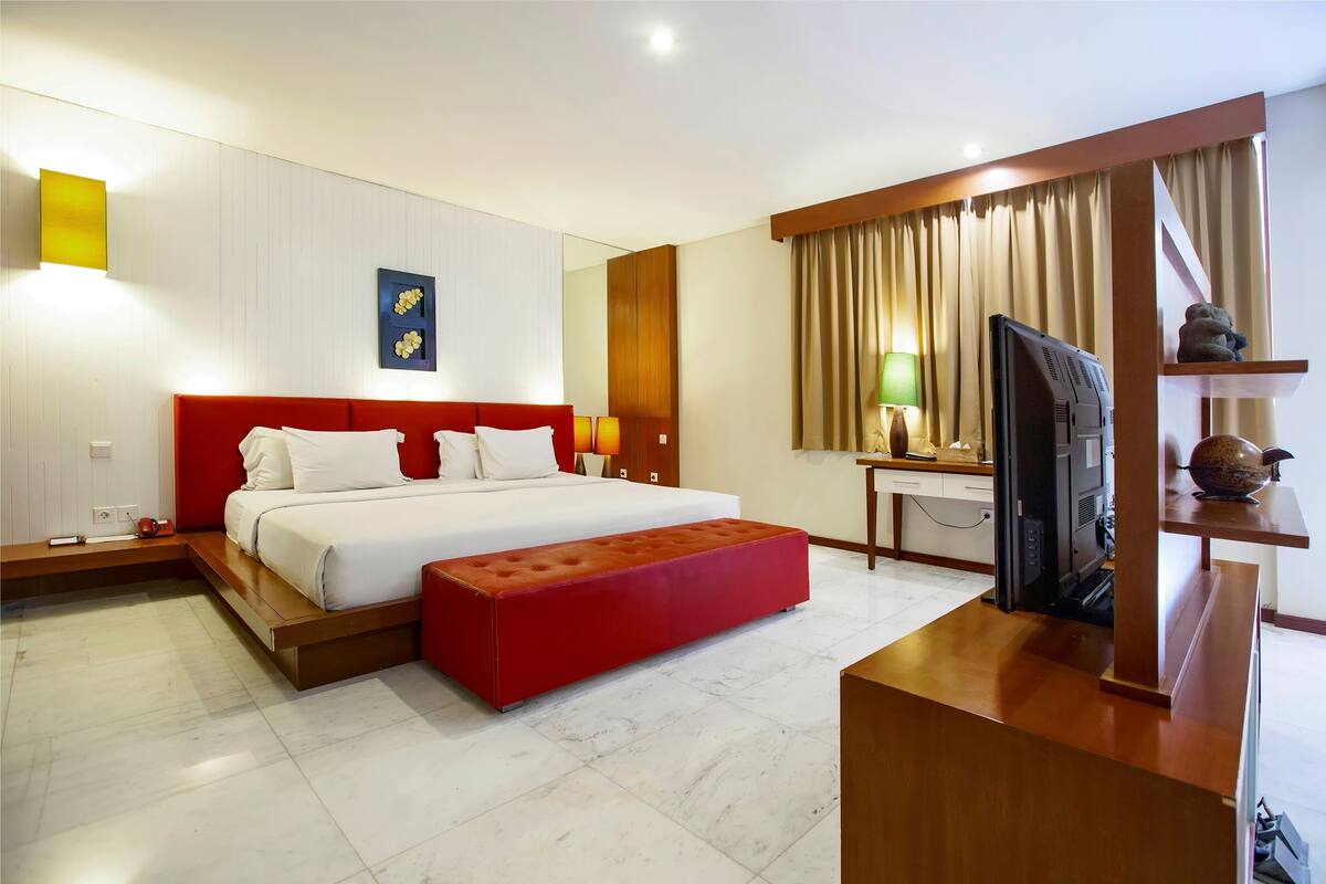 Quarto de hotel moderno com uma cama estofada vermelha, televisão de tela plana, escrivaninha com abajur e uma grande janela com cortinas.