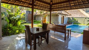A covered outdoor area with a pool, wooden furniture, and lush greenery. - Abi Bali Resort Villas & Spa (Jimbaran)
