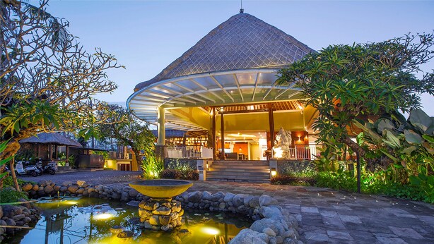 A modern building with a covered entrance, a pond with a fountain, and surrounding greenery. - Abi Bali Resort Villas & Spa (Jimbaran)