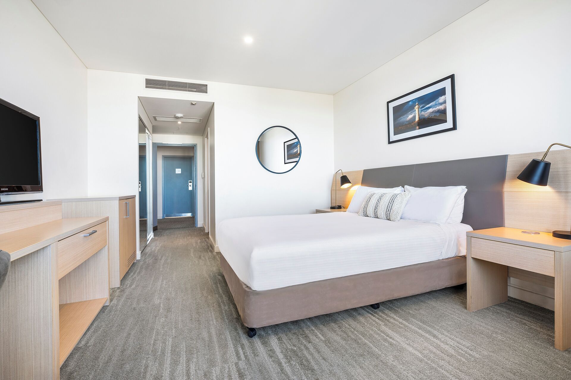 Quarto de hotel com cama, mesa de cabeceira, luminária, TV e um corredor que dá para outro quarto.