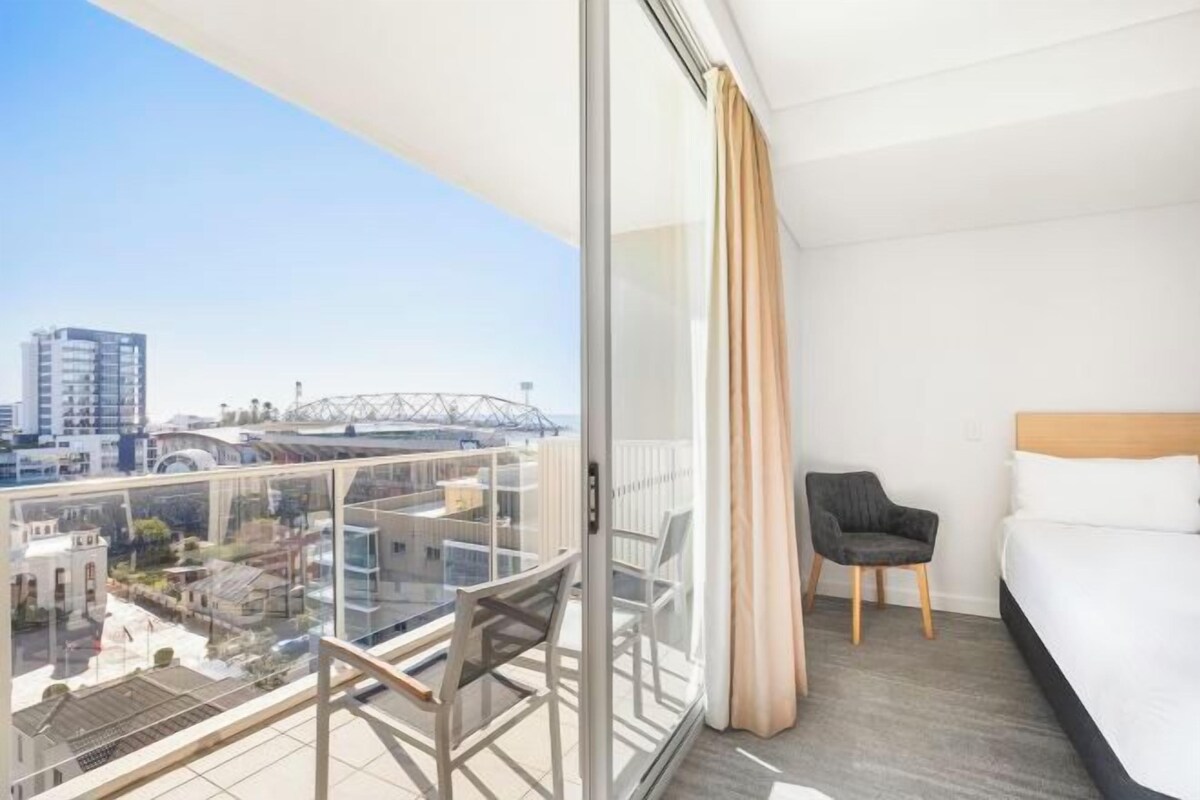 Habitación de hotel con balcón, cama, sillón y vista a la ciudad.