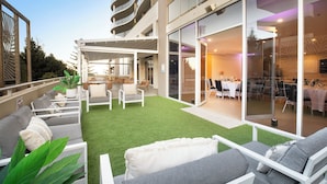 Un patio moderno al aire libre con mobiliario y una zona de comedor, conectado a un edificio con grandes puertas de cristal.