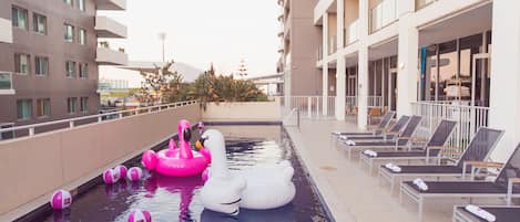 A rooftop pool with inflatable swan and flamingo floats, lounge chairs, and a building with balconies.