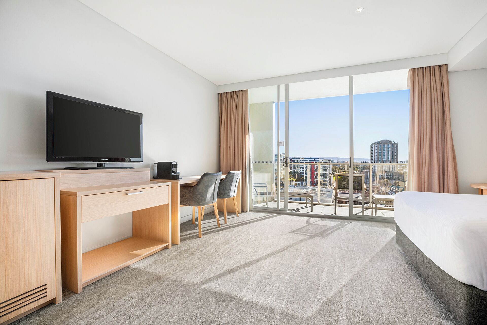 A hotel room with a large bed, a desk with a television, a chair, and a view of the city through large windows.