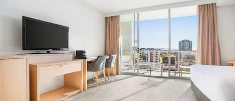 Habitación de hotel con una cama grande, un escritorio con televisión, una silla y vistas a la ciudad a través de ventanas amplias.