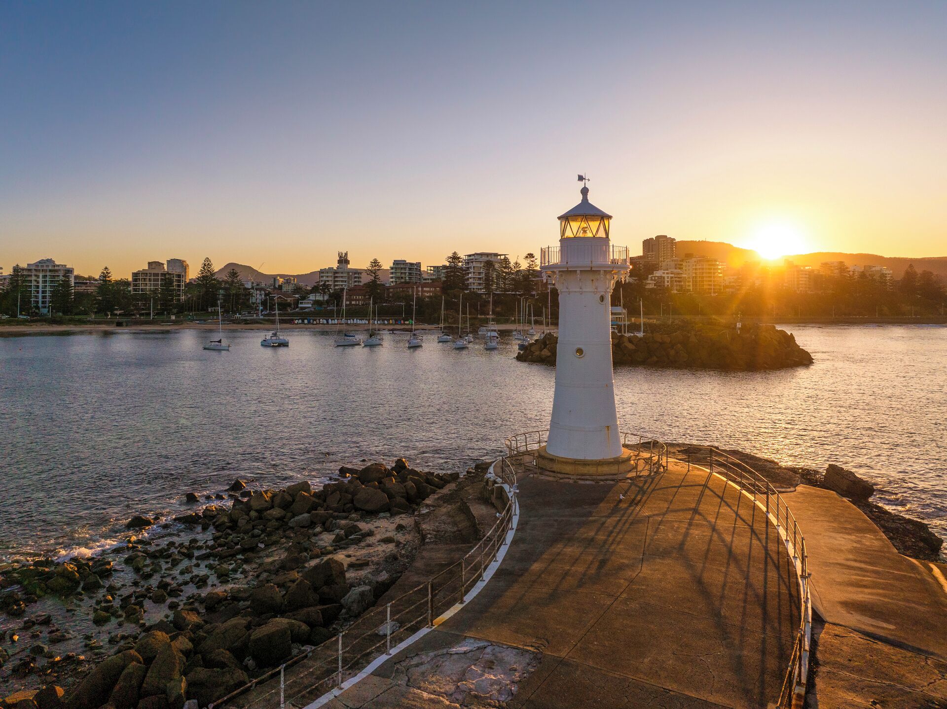 A lighthouse by a calm harbor with boats, a clear sky, and buildings in the background.