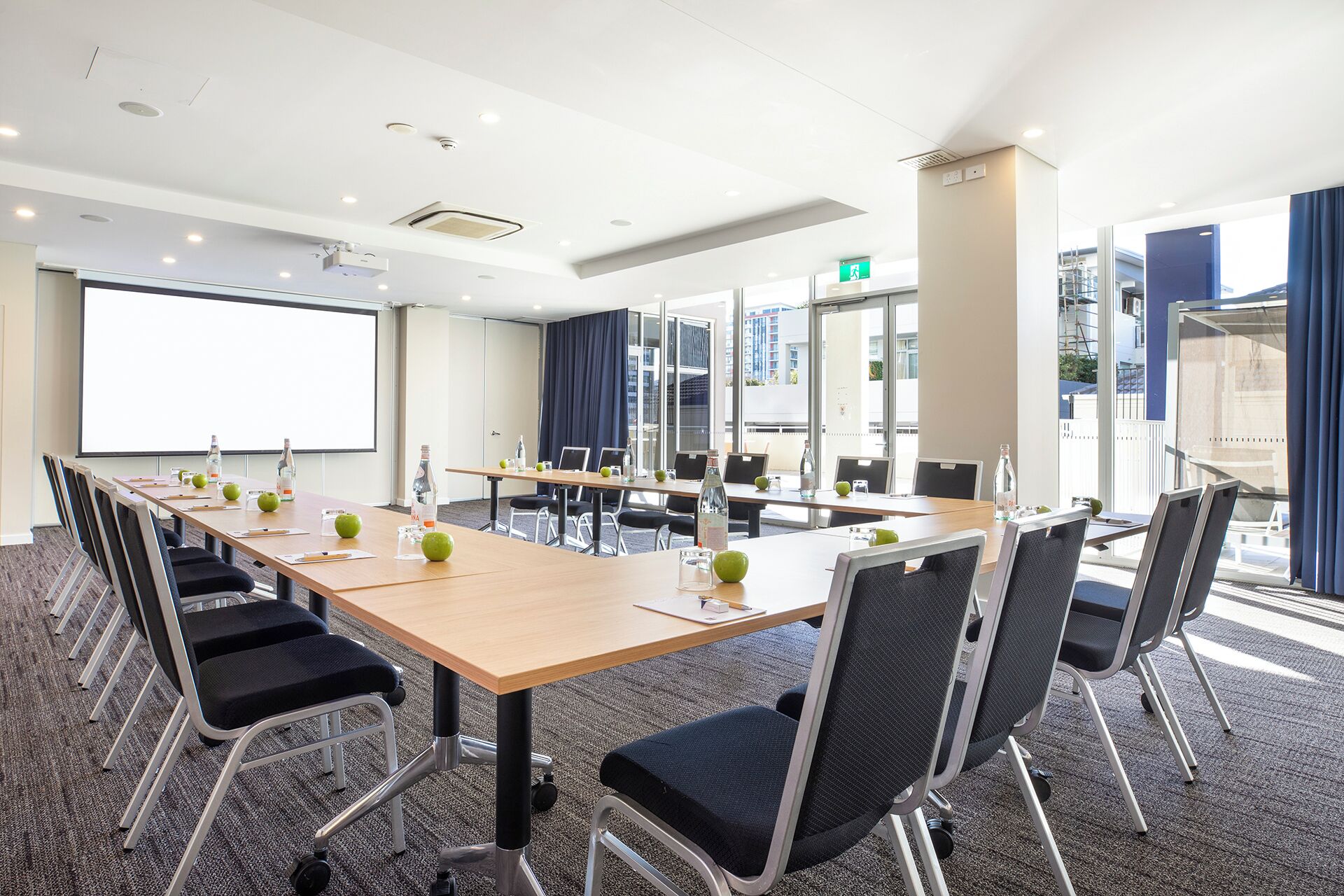 A conference room with a long table, chairs, a projector screen, and a large window with curtains.