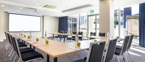 A conference room with a long table, chairs, a projector screen, and a large window with curtains.