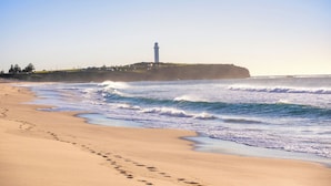 Una playa con un faro en un acantilado, olas rompiendo en la orilla y huellas en la arena.