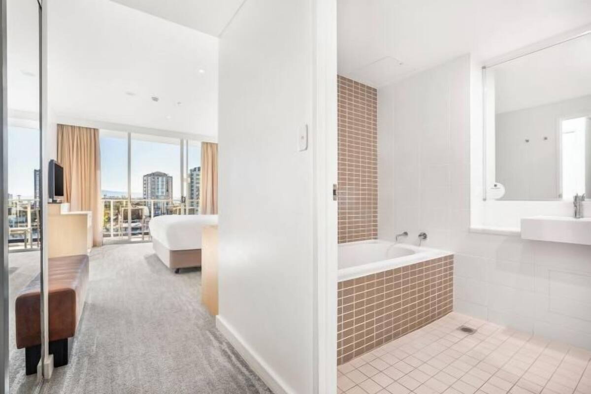 A modern hotel room with a city view, a large bed, a bathroom with a bathtub, and a balcony with a sofa.
