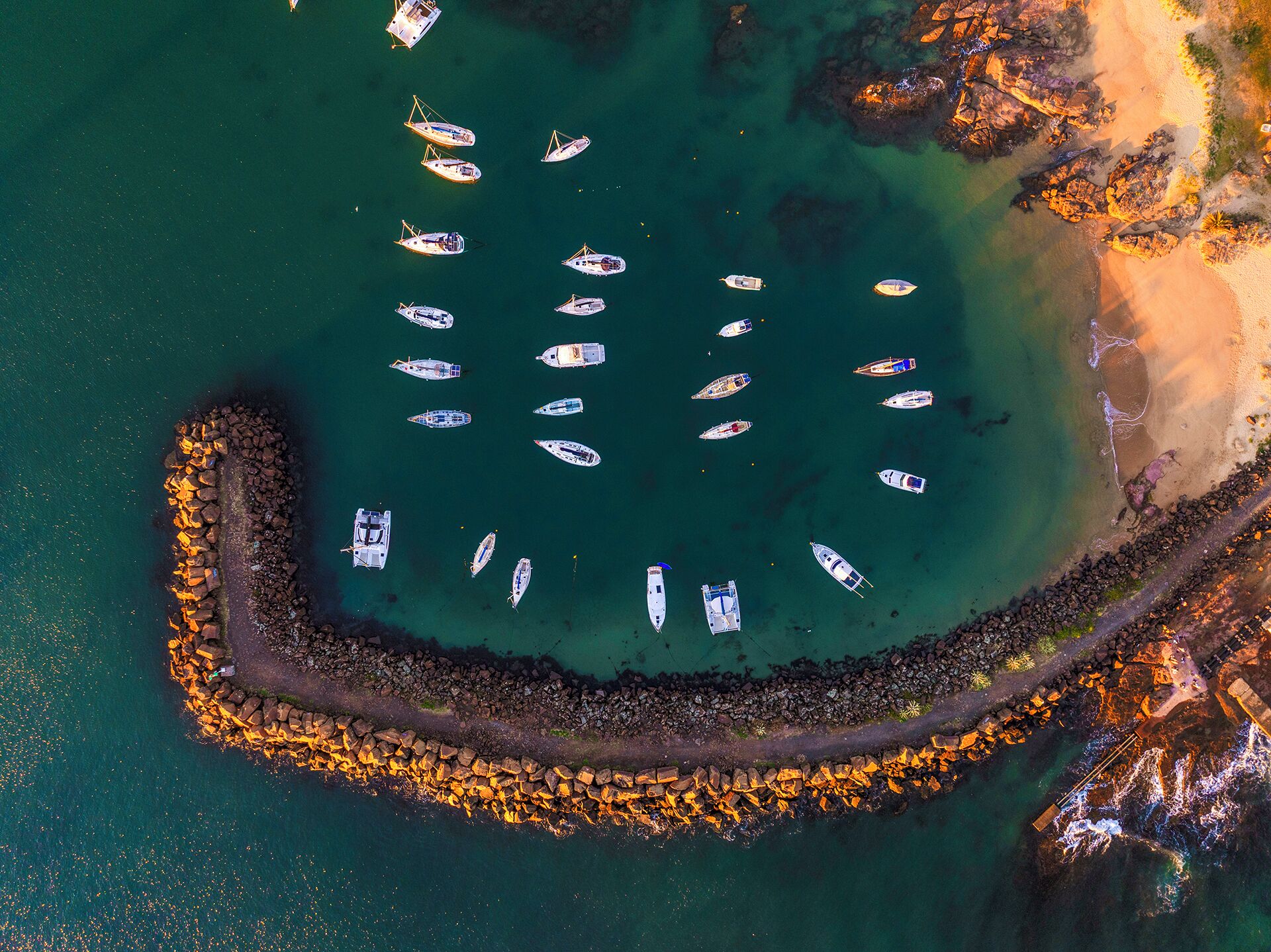An aerial view of a marina with boats docked and a rocky breakwater surrounding a sandy beach.