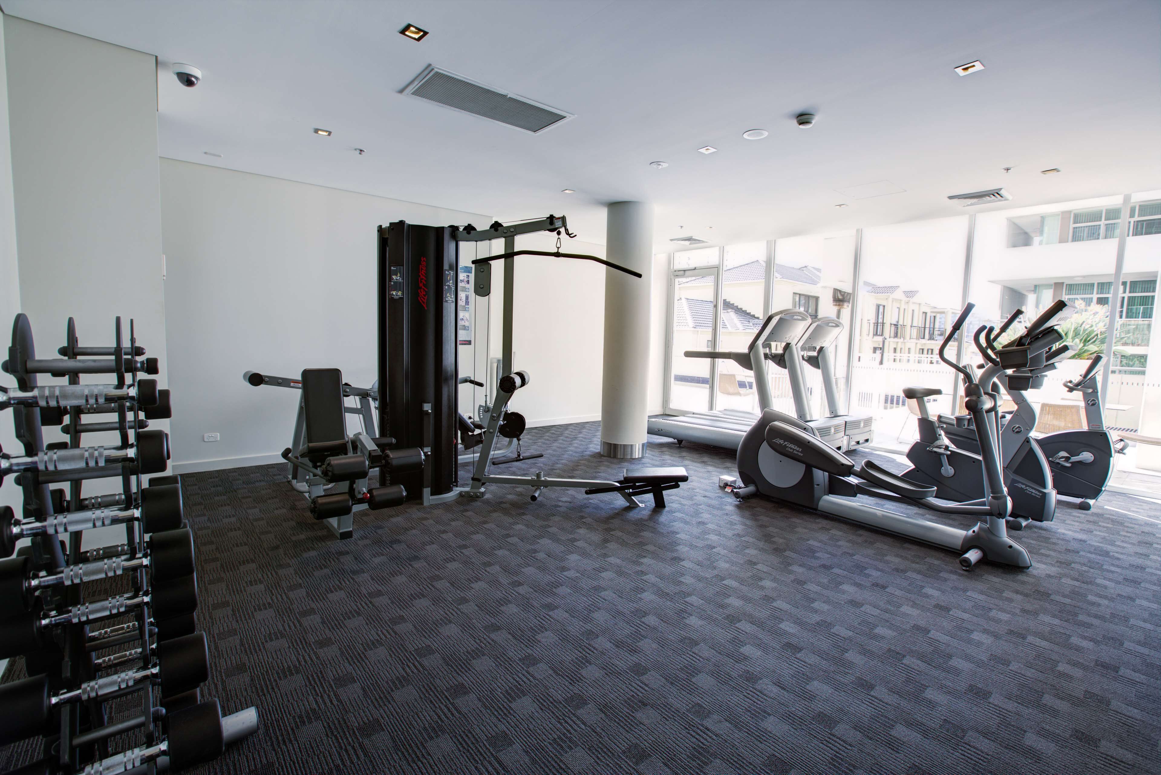 Een sportschool met diverse fitnessapparaten, grote ramen en een vloerbedekking.