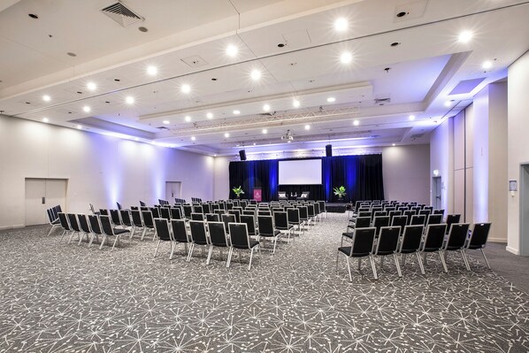 A conference room with rows of chairs, a stage with a projector, and a large screen.