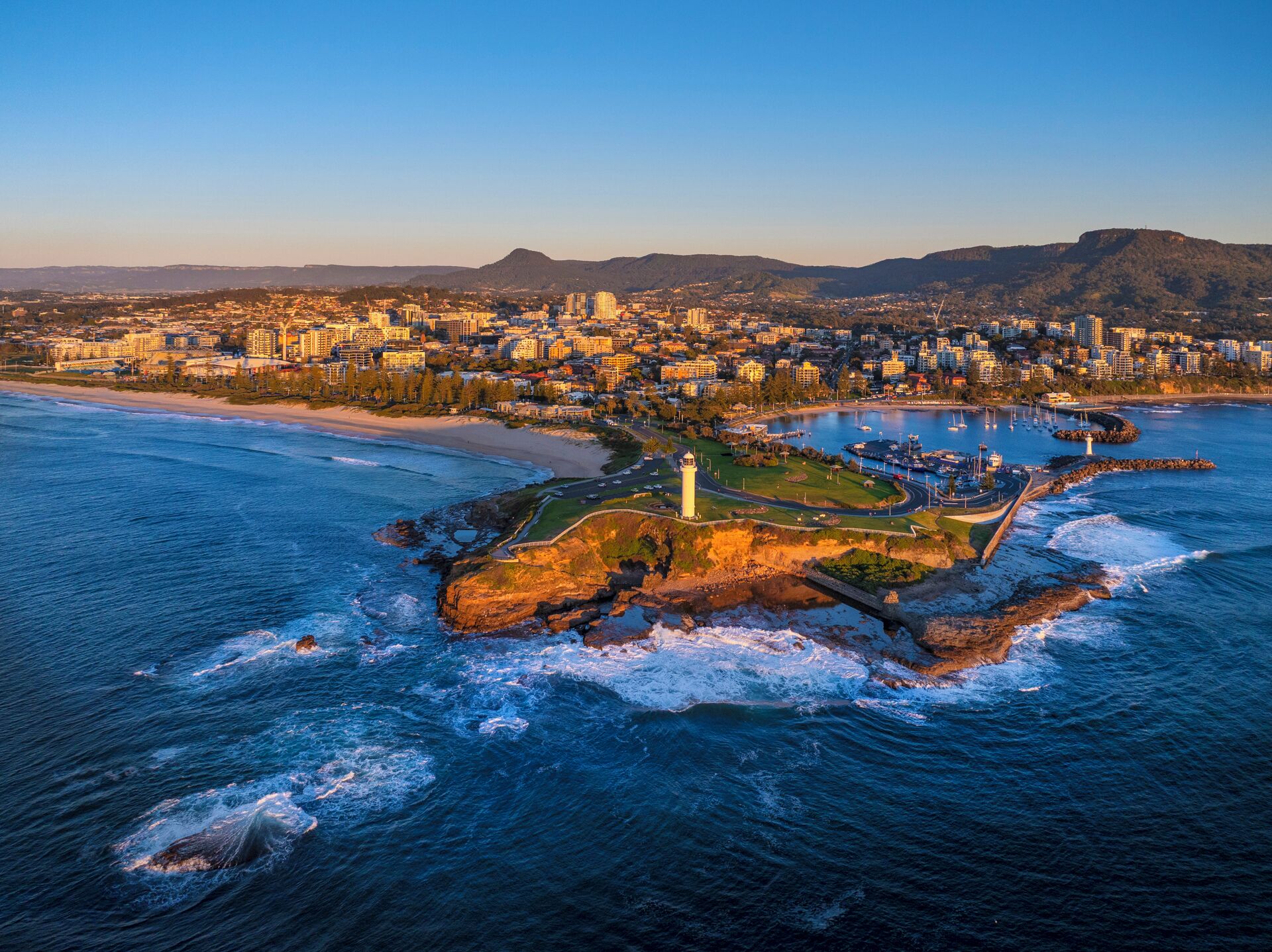 Vue aérienne d’une ville côtière, avec un phare imposant, une marina et une plage.
