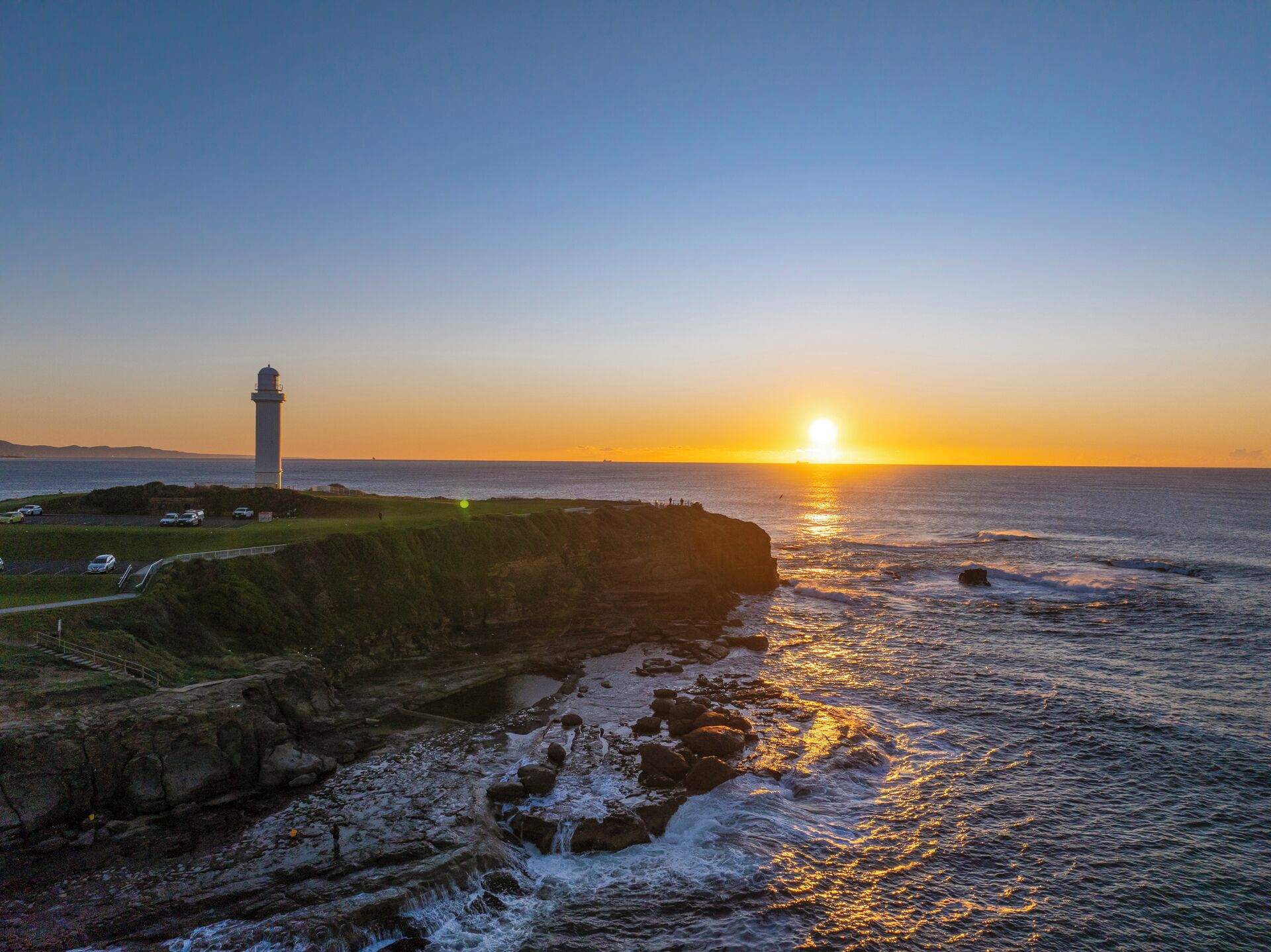 Paisaje costero con un faro, costa rocosa y un atardecer sobre el océano.