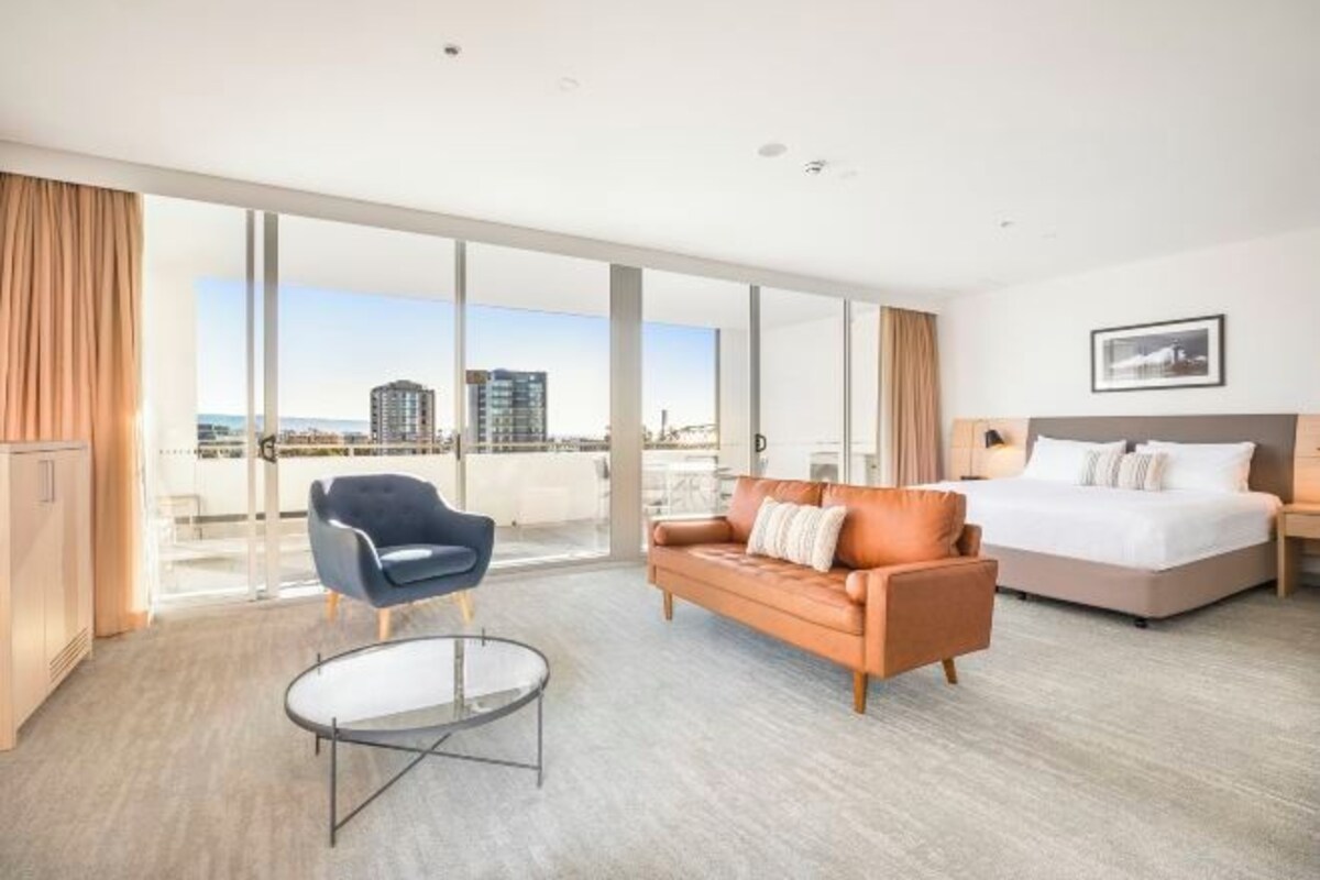 Habitación de hotel con cama, sofá, sillón, mesita de vidrio y vista a la ciudad a través de amplias ventanas.