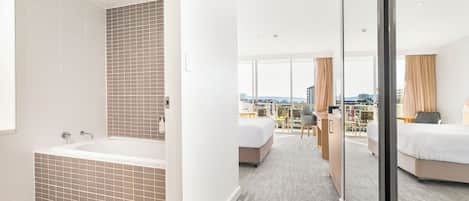 Une chambre d’hôtel moderne dotée d’une grande baignoire, d’un balcon offrant une vue sur la ville, ainsi que d’une chambre séparée avec un lit et un bureau.