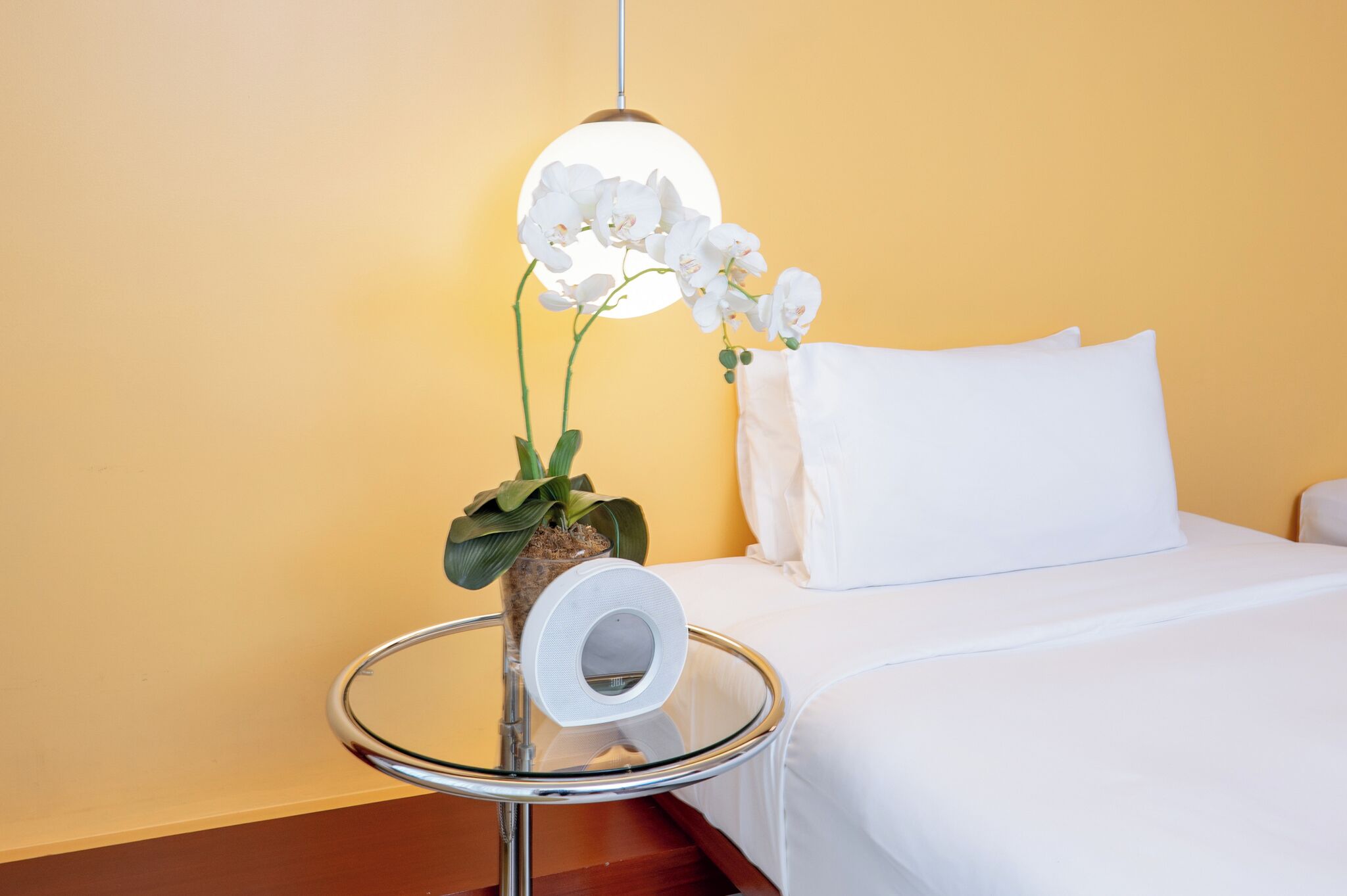 A hotel room with a bed, white pillow, a small round table with a glass top, a white speaker, and a potted plant with white flowers.