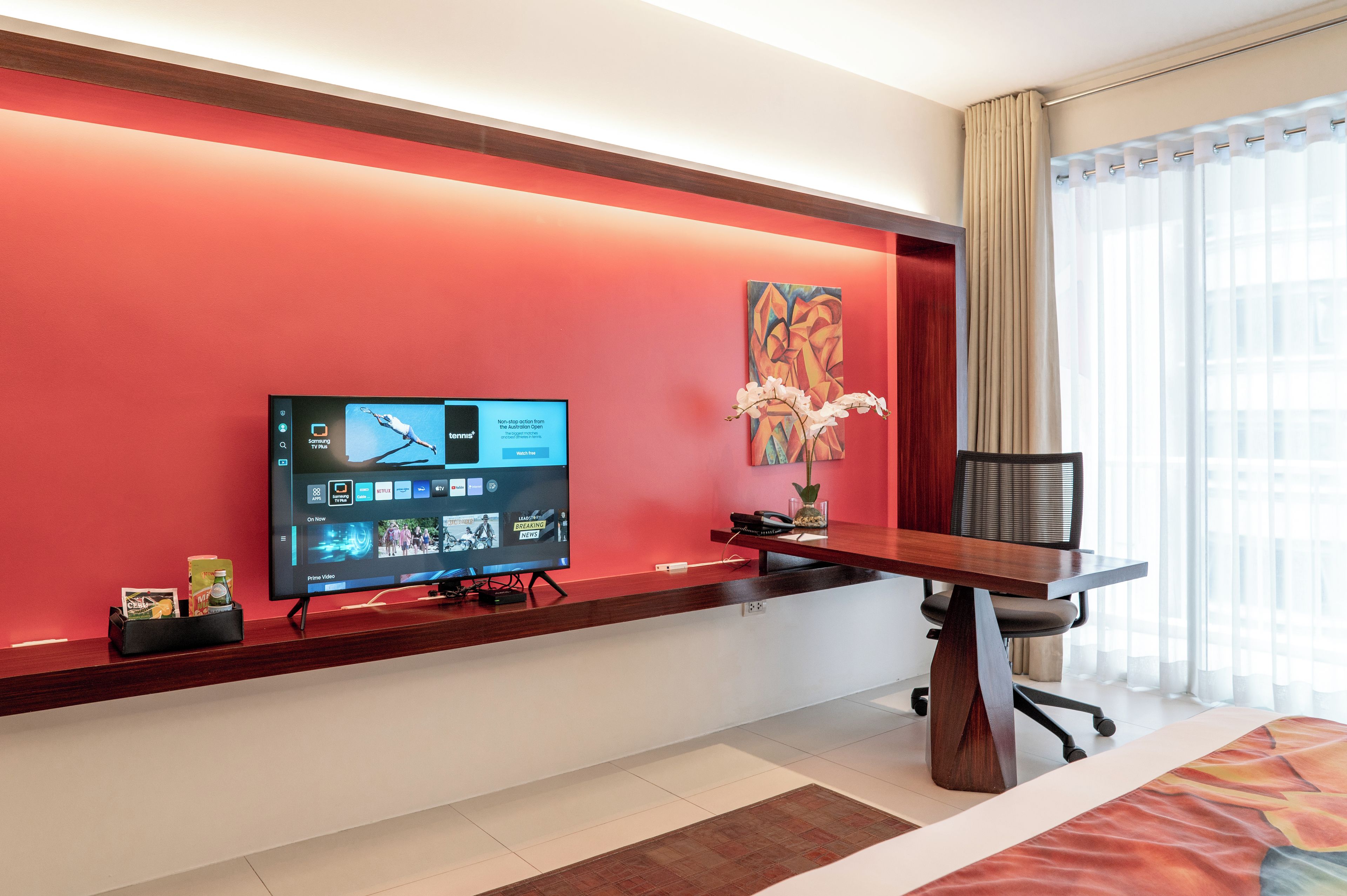 Een hotelkamer met een flatscreen televisie aan de muur, een bureau met stoel, een vaas met bloemen en een bed met een rood-oranje beddegoed.