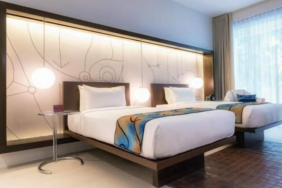 Une chambre d’hôtel au design moderne, dotée d’un grand lit avec une tête de lit intégrant un luminaire, d’une table de chevet et offrant une vue sur le paysage urbain grâce à de grandes fenêtres.