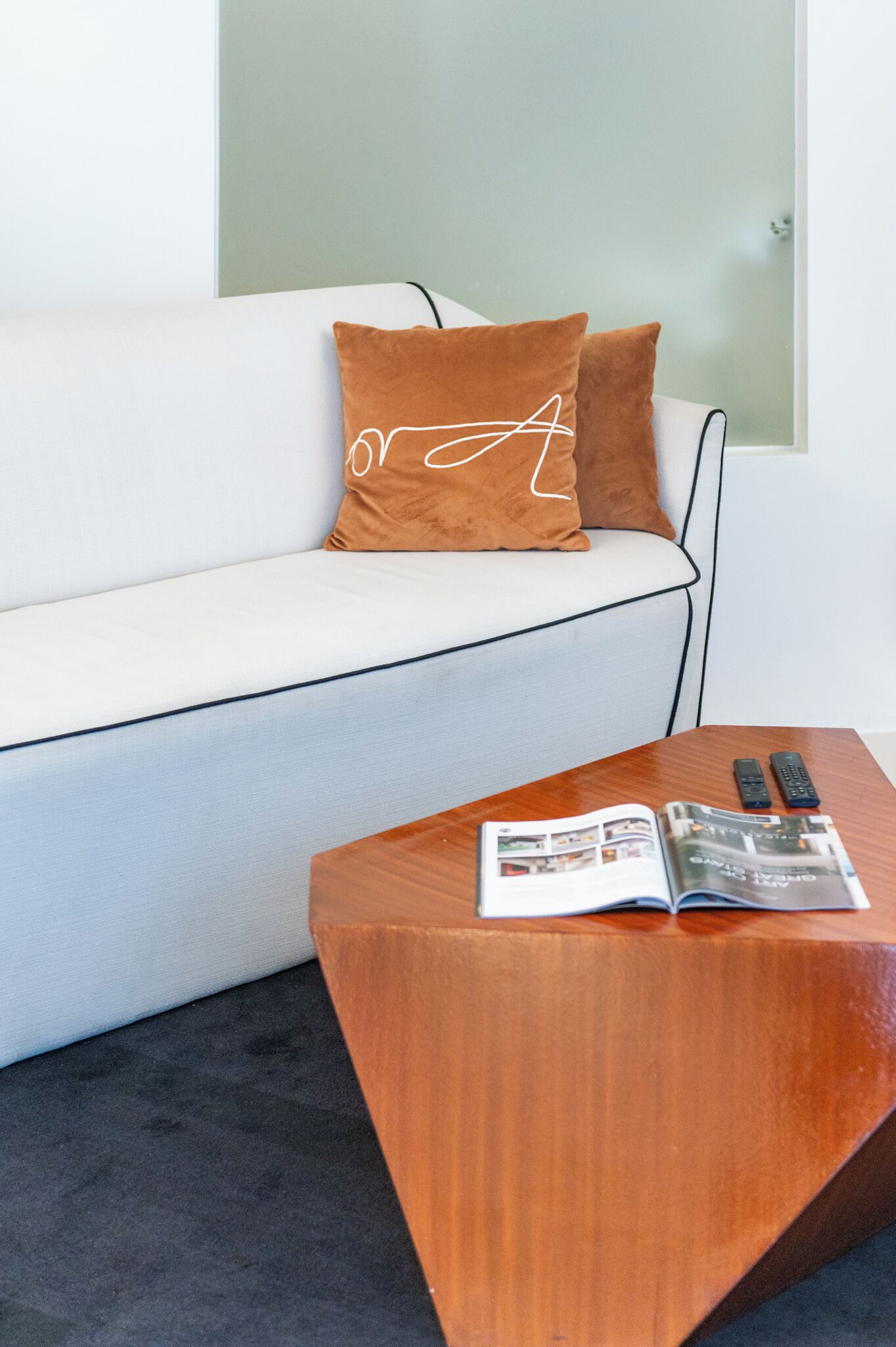 A modern living room with a white sofa, two brown pillows, and a wooden coffee table with a magazine and remotes.
