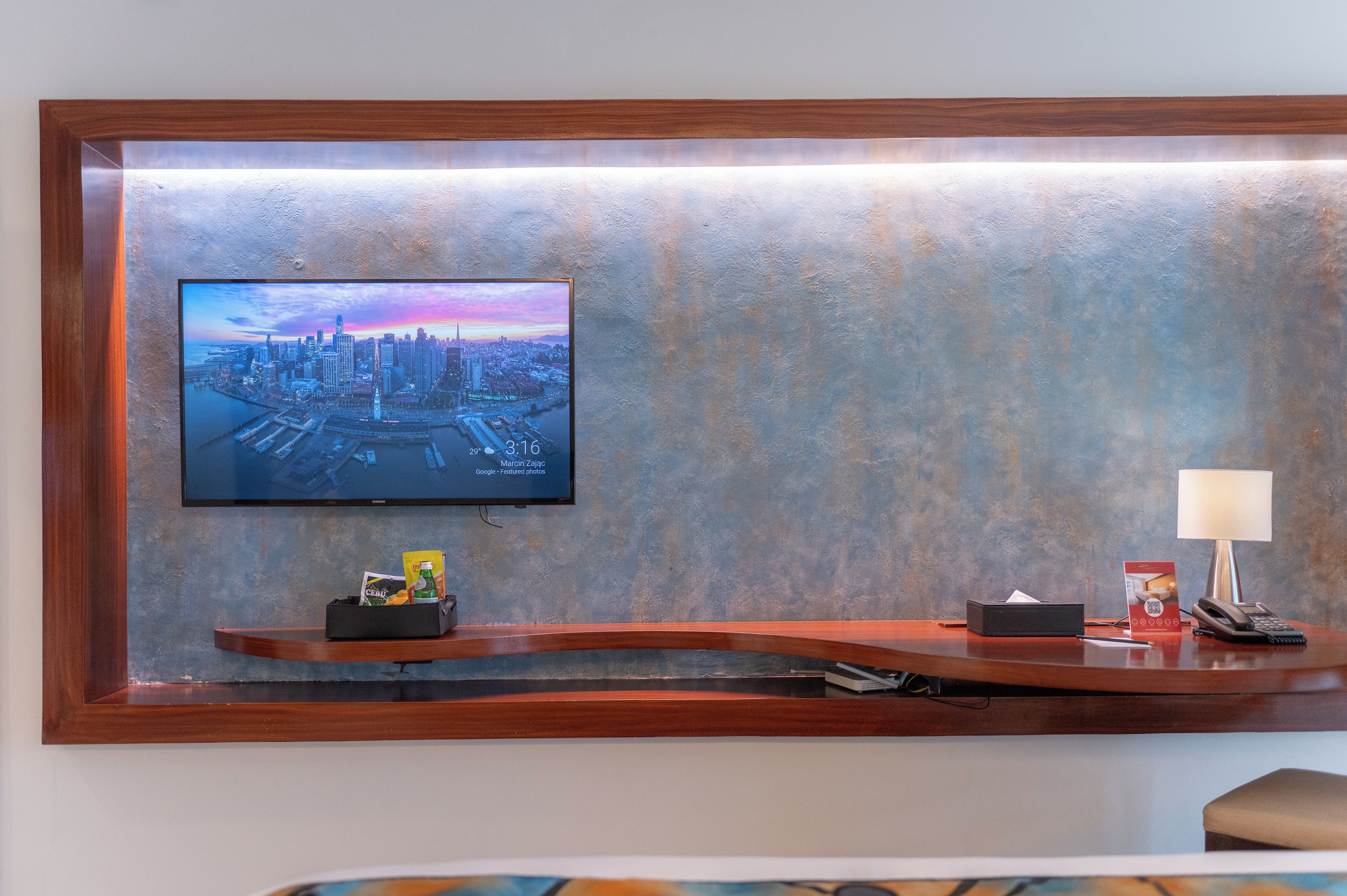 A hotel reception area with a mounted television displaying a cityscape, a desk with a telephone, a lamp, and a small seating area.