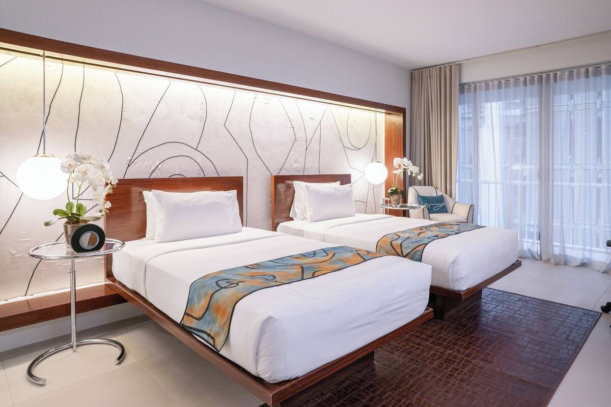 Une chambre d’hôtel avec deux lits, une table de chevet avec une lampe, une chaise et une grande fenêtre avec des rideaux.