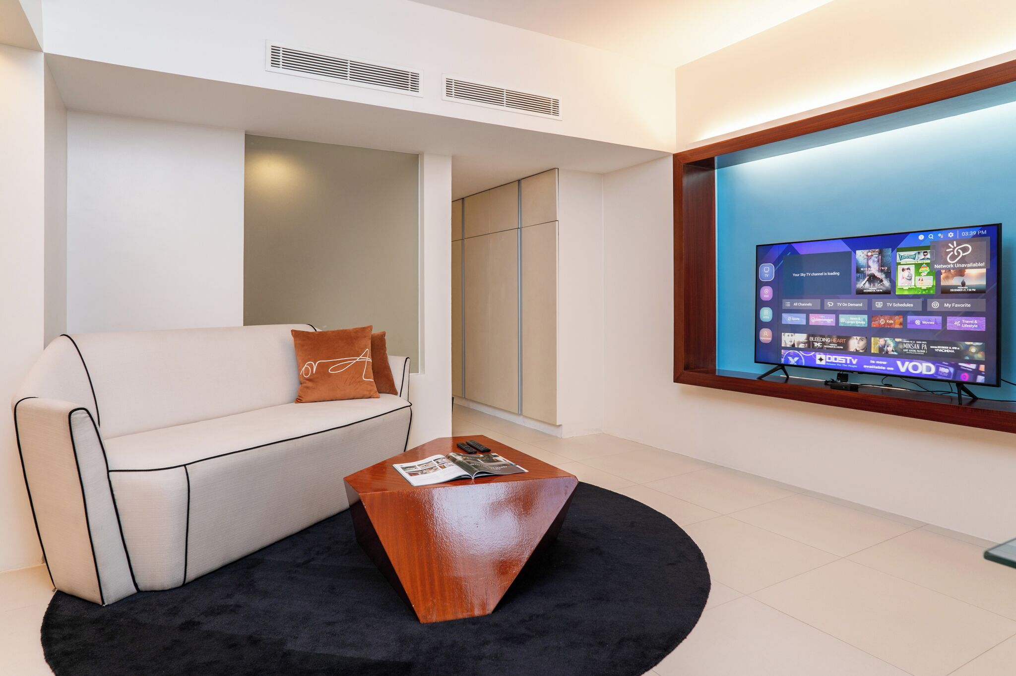 Una sala de estar moderna con un sofá blanco, un tapete negro y una mesa de centro de madera. Un televisor de pantalla plana está montado en la pared mostrando varios canales.