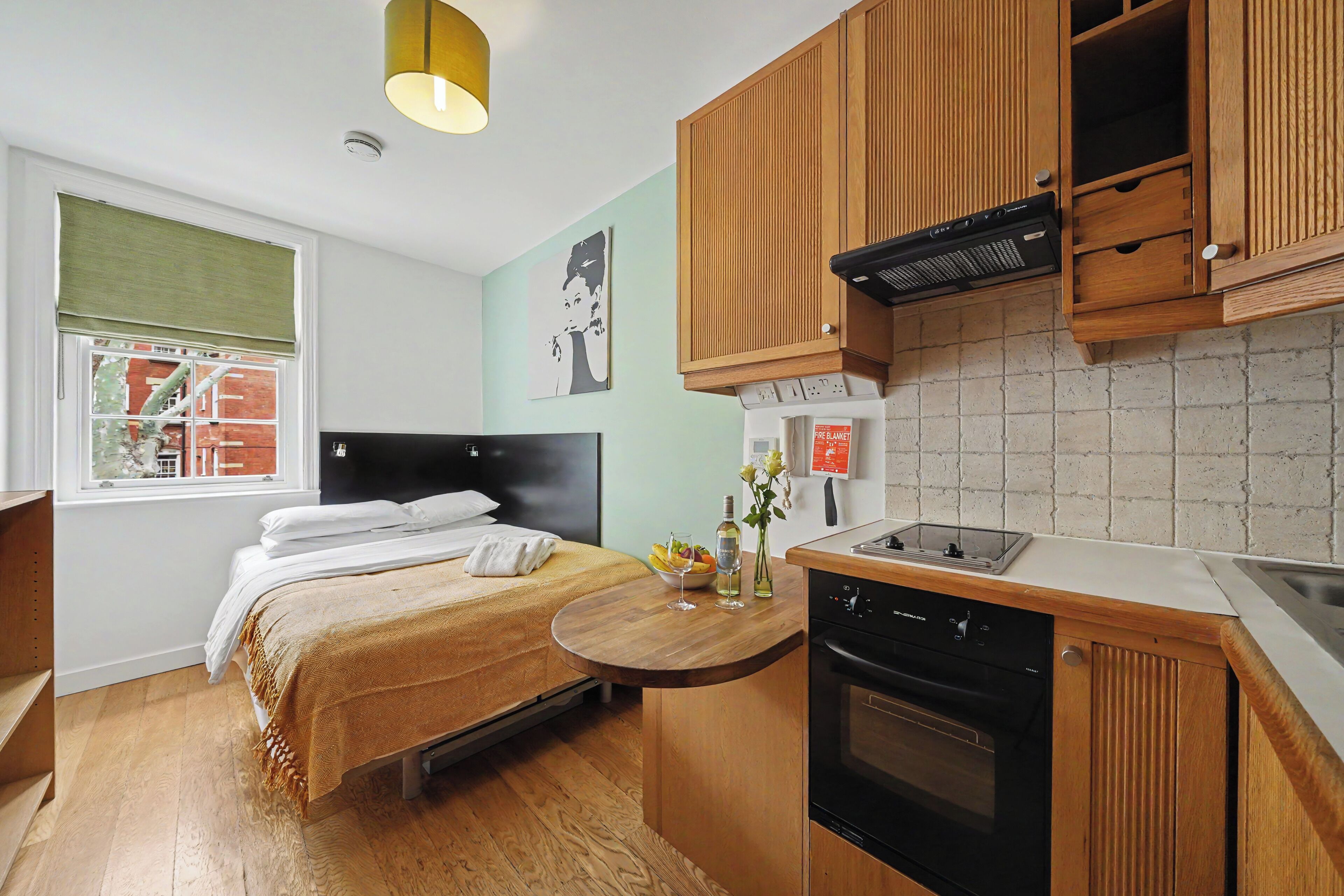 A compact hotel room with a bed, a small dining table, a kitchen area with a stove and dishwasher, and a window with a view of buildings.