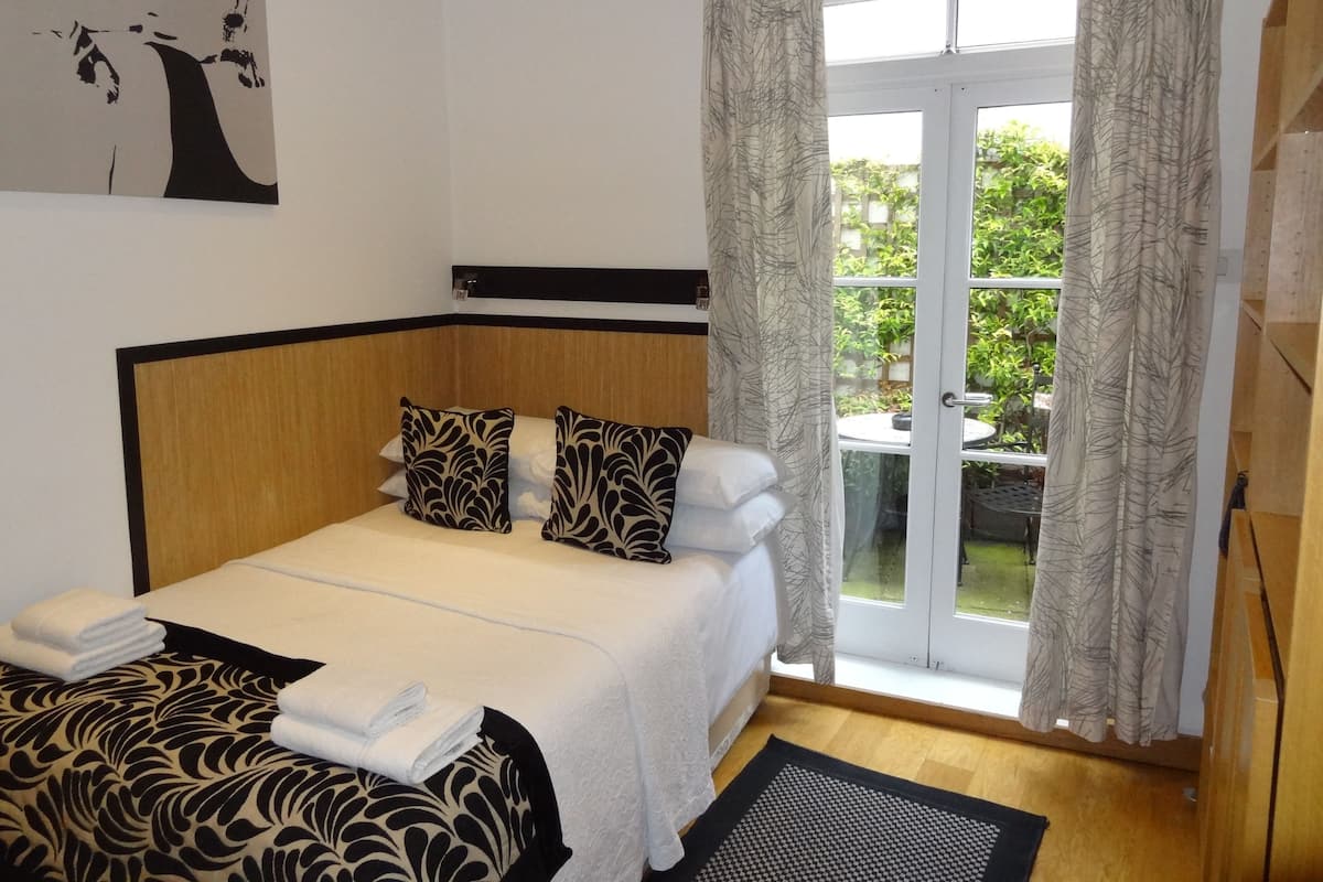 A hotel room with a bed, a window with curtains, a view of greenery outside, and a painting on the wall.
