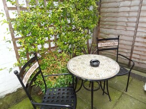 Uma pequena área externa com mesas e cadeiras, cercada por um muro de madeira e vegetação.